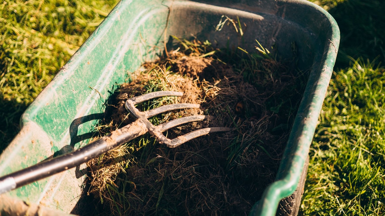 A pitchfork lies in a green wheelbarrow filled with straw and organic matter.