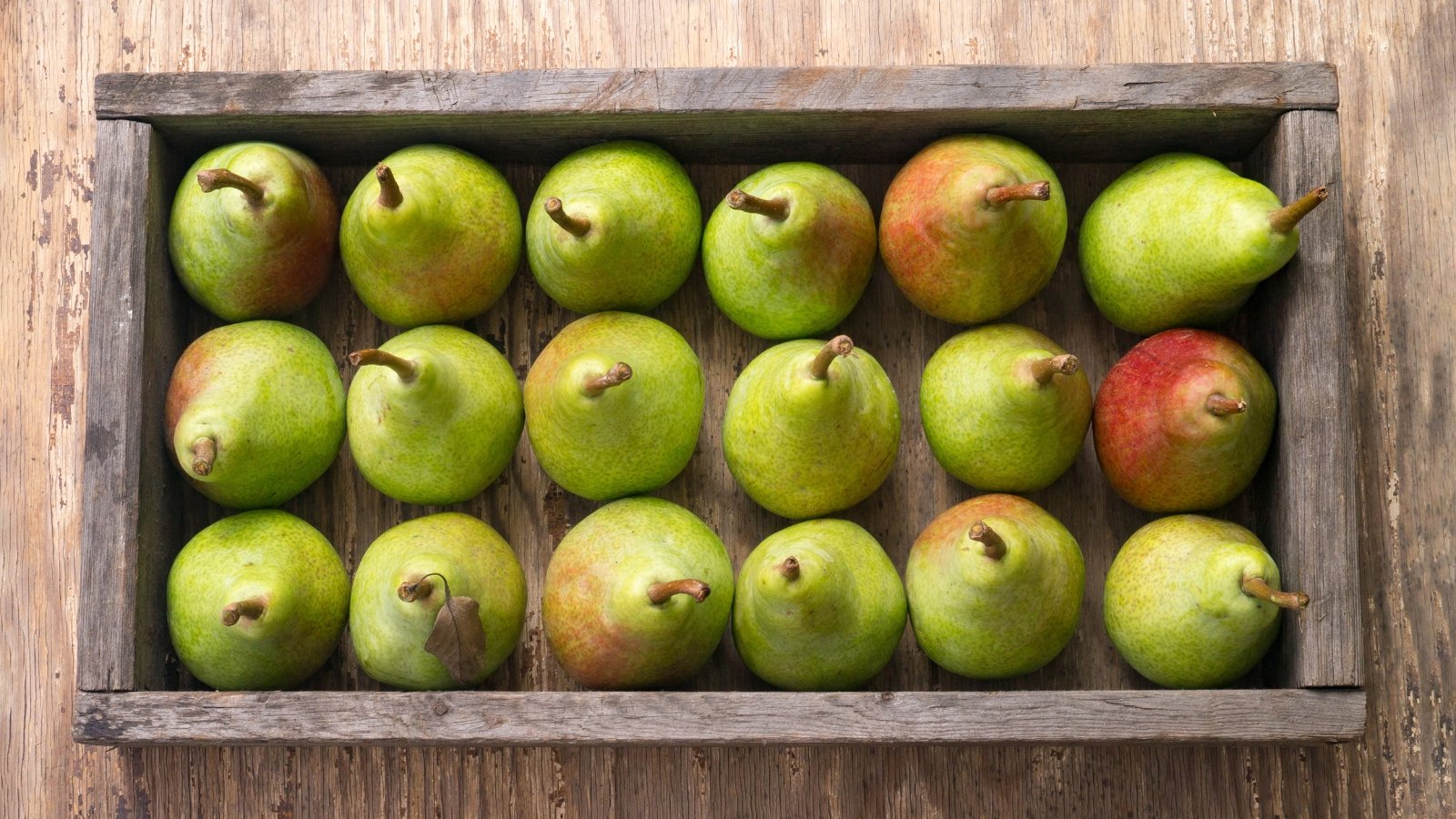 A rustic wooden crate displaying an orderly arrangement of green fruits, some tinged with red blush near the stems, their smooth surfaces glowing under soft natural light, with a rough wooden surface underneath.